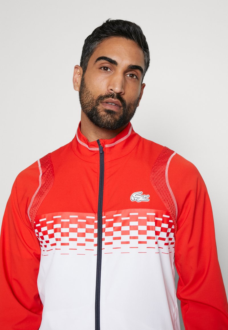 Uomo con barba curata che indossa una giacca a zip rossa e bianca con un motivo a quadri e un piccolo logo di coccodrillo.