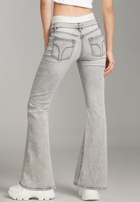 Gray flared jeans with a high waistband, back pockets featuring a distinctive pattern, and a raw hem. Paired with white chunky sneakers.