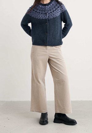 Personne portant un cardigan bleu foncé à motifs boutonné, un pantalon large en velours côtelé beige et des bottines noires, debout sur un sol gris.