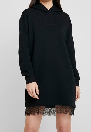 Femme portant une robe à capuche noire à manches longues avec un bord en dentelle qui apparaît à l'ourlet, debout devant un fond uni clair.