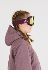 Skibril met een kastanjebruine band, zwarte schuimrubber padding en reflecterende gele lenzen. Draagt gedragen door iemand in een paarse jas met een capuchon.