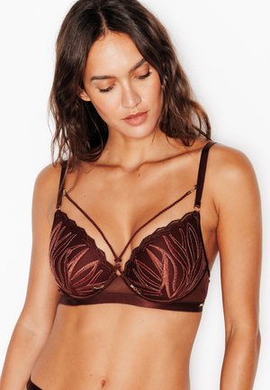 Femme aux longs cheveux bruns portant un soutien-gorge transparent marron foncé avec des motifs de feuilles brodés complexes et des fines bretelles décoratives.