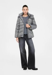 Siva pufer jakna sjajnog završetka, zatvaranje na patentni zatvarač, odvojiva kapuljača i vodoravni prošivani uzorak. Uparena s tamnim džemperom i širokim trapericama.