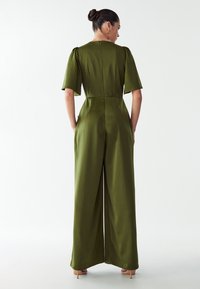 Ženska v olivno zeleni kratkolasni široki kombinaciji z zadrgo na hrbtu, ki stoji ob belem ozadju.