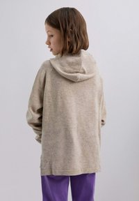 Hellbeige Kapuzensweatshirt mit weicher Textur, lässiger Passform und abgesenkten Schultern, kombiniert mit lila Hosen.