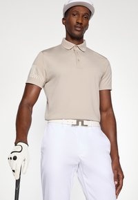 Beige polo skjorte med korte ermer, teksturerte detaljer på skuldrene. Kombinert med hvite bukser og et hvitt belte med metallspenne. Golfhanske i hånden.
