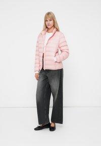 Giacca trapuntata rosa chiaro con cuciture orizzontali e chiusura zip, abbinata a jeans grigi larghi e ballerine nere, su uno sfondo bianco.