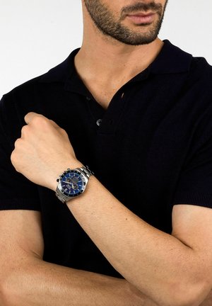 Orologio in acciaio inossidabile argentato con quadrante blu, accenti argentati e funzione cronografo. Indossato su un polso con tonalità di pelle chiara.