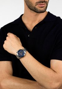 Orologio in acciaio inossidabile argentato con quadrante blu, accenti argentati e funzione cronografo. Indossato su un polso con tonalità di pelle chiara.
