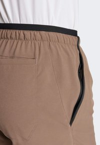 Brązowe sportowe spodenki z elastycznym czarnym pasem, boczną kieszenią na zamek i gładką fakturą materiału. Posiadają dyskretną tylną kieszeń.