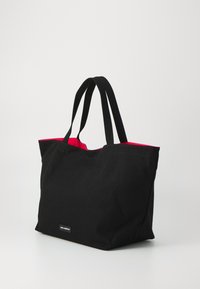 Sac fourre-tout noir en toile avec intérieur rouge, doté de deux poignées noires et d'une petite étiquette de logo en bas. Forme rectangulaire, design simple.