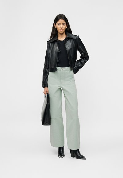 Γυναίκα που φοράει μαύρο δερμάτινο μπουφάν, μαύρο τοπ, ανοιχτοπράσινο παντελόνι με φαρδιά μπατζάκια, μαύρα μποτάκια και κρατάει μαύρη τσάντα, στέκεται σε λευκό φόντο.