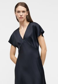 Γυναίκα με ίσια καστανά μαλλιά, φοράει μαύρο σατέν φόρεμα με βαθύ V και κοντά μανίκια τύπου φτεροκοπήματος, μπροστά σε απλό ανοιχτό φόντο.