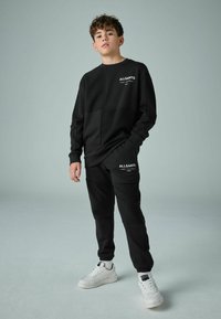 Svart träningskläder set som innehåller en långärmad topp med rund hals och matchande joggers. Båda plaggen har vita logotillbehör.