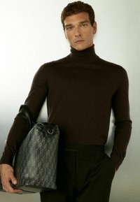 Dunkelbrauner Rollkragenpullover, taillierte Passform. Das Modell hält eine strukturierte schwarze Tasche mit geometrischem Muster und Lederakzenten.