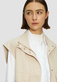 Veste sans manches beige avec boutons-pression et fermeture éclair, portée sur une chemise blanche à col. Tissu lisse avec un design décontracté.