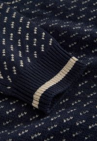 Pull en maille marine avec des motifs en zigzag crème. Présente une finition texturée et un poignet côtelé avec une bande crème. Matière en laine douce.