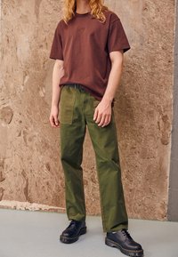 Hnědé bavlněné tričko s malým logem na hrudi, spojené se zelenými bavlněnými kalhotami. Černé šněrovací boty s tlustou podrážkou.