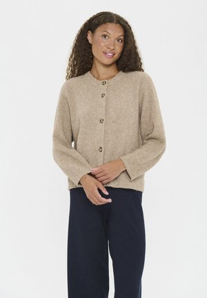 Woman with curly hair wearing a beige buttoned cardigan and navy wide-leg pants, standing with hands loosely clasped.
