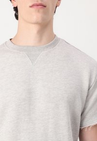 Tricou din bumbac gri deschis, cu guler rotund, mâneci scurte și marginile brut. Materialul are o textură moale, cu detalii de cusătură minime.