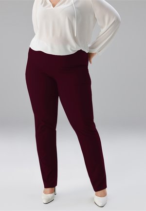 Frau trägt dunkelburgunderfarbene, eng anliegende Hose, weiße Bluse und weiße spitze Pumps, steht vor einem schlichten grauen Hintergrund.