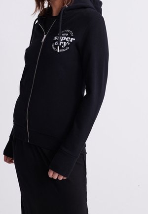 Kobieta ubrana w czarną bluzę z zamkiem, z białym, okrągłym logo Superdry na lewej piersi, w połączeniu z czarnymi spodniami lub spódnicą.