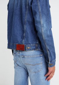 Denimjakke i mørkeblå med et lærmerke i livet, sammen med lyseblå denimjeans med baklommer og synlige sømmer.