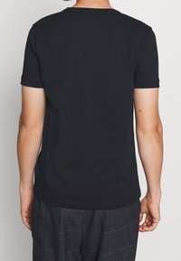 Osoba v jednoduchém černém tričku s krátkým rukávem a tmavě modrých kostkovaných kalhotách, stojící zády k fotoaparátu na bílém pozadí.