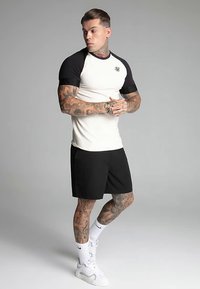 Vit och svart kortärmad sporttröja, svarta shorts, med synliga tatueringar på armarna och bär vita sneakers med svarta detaljer.