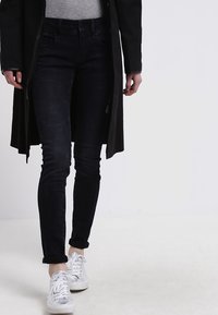 Pantalones vaqueros negros de corte ajustado, con una cintura de altura media y un ligero desgaste. Combinados con zapatillas blancas y una blusa de punto gris bajo un abrigo negro.