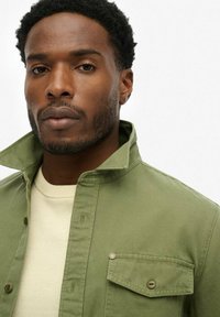 Groene button-up jas met een spits kraag, borstzak en een gestructureerde afwerking, gelaagd over een crèmekleurige trui.