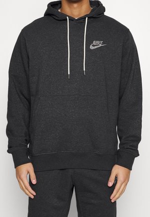 Man wearing dark gray Nike hoodie with white drawstrings and matching dark gray pants, standing with hands relaxed at sides.