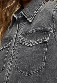 Nahaufnahme einer dunkelgrauen Jeansjacke mit aufgenähten Klappentaschen und metallenen Druckknöpfen, die über einem Hemd getragen wird, mit langem Haar, das sichtbar ist.