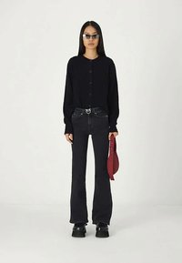 Kvinna klädd i en svart knäppt kofta, svarta utsvängda jeans, svarta plattformsskor, solglasögon, och håller en liten röd handväska.