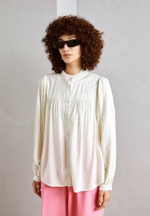 Weiße Bluse mit Knöpfen und Rüschensaum, gesammelten Details an der Brust und langen Ärmeln. Kombiniert mit pinkfarbenen weiten Hosen.