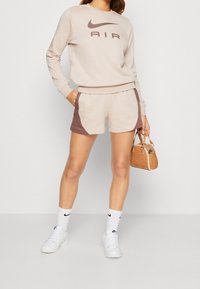 Beżowa bluza Nike z logo "AIR", zestawiona z beżowo-brązowymi szortami w stylu color-block. Białe skarpetki i trampki, trzymająca małą brązową torebkę.