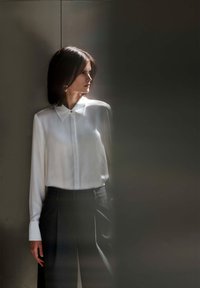 Biela hodvábna košeľa s golierom a detailmi na gombíkoch, kombinovaná s tmavými upravenými nohavicami. Mäkká textúra a elegantné strihanie na neutrálnom pozadí.