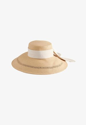 Platas rāmas, bēša krāsas salmu saules cepure ar vieglu auduma joslu, kas aizmugurē sasieta lokā, un dekoratīvām caurumiem pie malas.