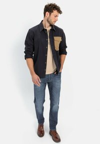 Donkerblauwe overhemdenjack met een beige borstzak, gedragen over een beige shirt. Gecombineerd met blauwe jeans en bruine lederen schoenen.