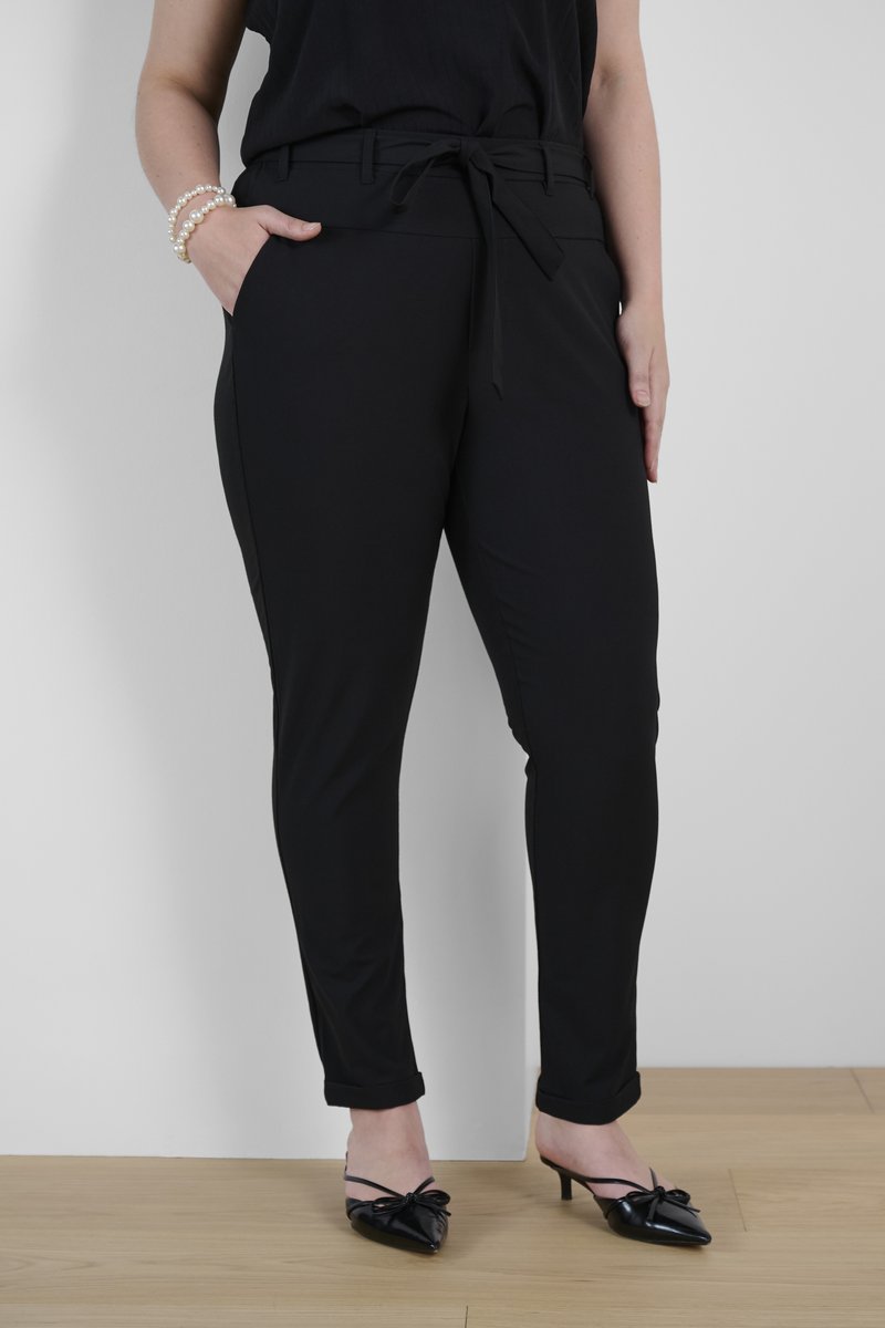 Schwarze maßgeschneiderte Hose mit einem Taillengürtel, seitlichen Taschen und schlankem Schnitt. Glatte Textur mit umgeschlagenen Säumen; kombiniert mit schwarzen spitzen Absatzschuhen.