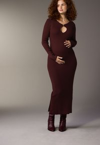 Bordeauxfarbenes, langärmeliges Kleid aus Rippstrick mit Ausschnitt in Tropfenform, figurbetonter Silhouette, seitlichem Schlitz und kombiniert mit knielangen bordeauxfarbenen Stiefeln.