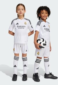 Dvoje djece nose bijele fudbalske komplete s crnim detaljima i Adidas logotipom. Dječak drži crno-bijelu fudbalsku loptu.