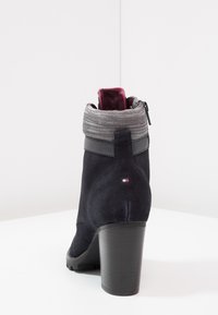 Námořnicky modrá semišová kotníková bota se šedým pleteným límcem, bočním zipem a robustním černým podpatkem. Na zadní straně je malý logo detail.