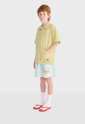 Niño pelirrojo con camisa de manga corta amarilla a cuadros, pantalones cortos azul claro con texto, calcetines blancos y chanclas rojas, de pie de lado.