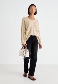 Maglione beige a trecce con scollatura a V, jeans neri a gamba dritta, scarpe con piattaforma marroni e una piccola borsa bianca con una sciarpa a fantasia.
