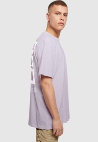 Lilla T-shirt med korte ærmer, der har fed hvid tekst og grafik, afslappet pasform, rund halsudskæring og blødt bomuldsstof.
