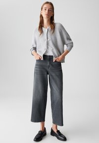 Femme portant un cardigan gris à boutons, un haut blanc, un jean noir délavé à jambes larges et des mocassins noirs, se tenant avec les mains dans les poches.