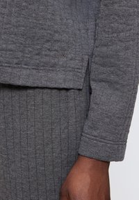 Grauer, strukturierter Stoff-Oberteil mit geripptem Design und einem Bündchenärmel. Enthält ein kleines, gesticktes Logo am Saum.