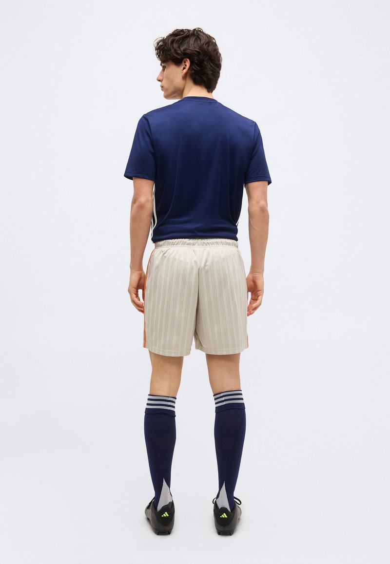 Hombre de pie de espaldas, vestido con una camiseta azul marino, pantalones cortos de rayas claras con los lados naranjas, calcetas altas hasta la rodilla de color azul marino y zapatos de fútbol negros.