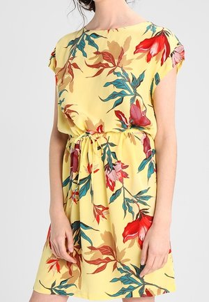 Robe fleurie jaune avec des feuilles rouges, roses et bleues, manches courtes, taille cintrée avec cordon, portée par une personne debout.